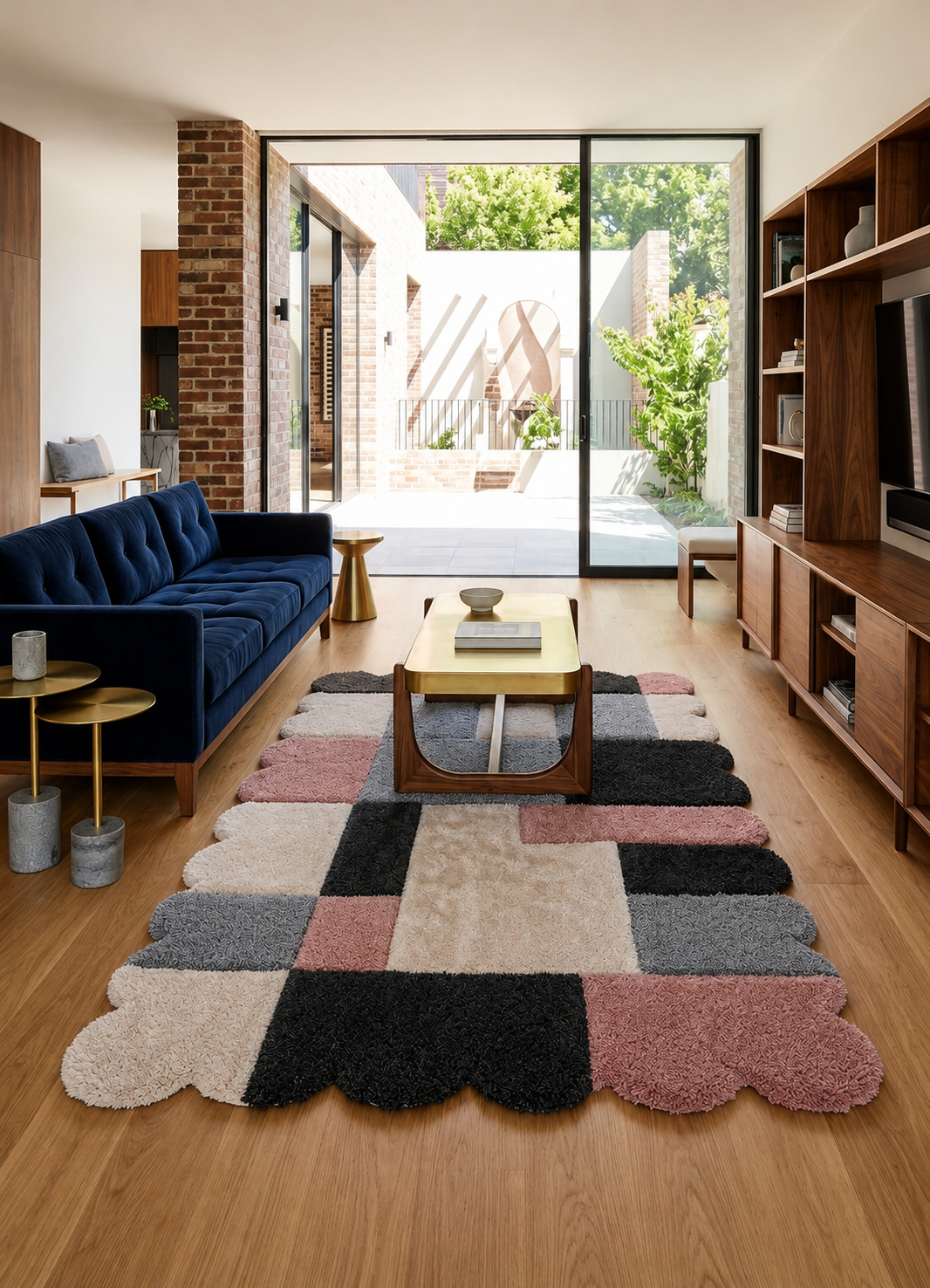 A modern living room featuring a large, scalloped patchwork area rug with plush shag texture in black, beige, pink, and gray. The room is styled with a deep blue velvet sofa, wooden shelving, and a glass sliding door leading to an outdoor patio.