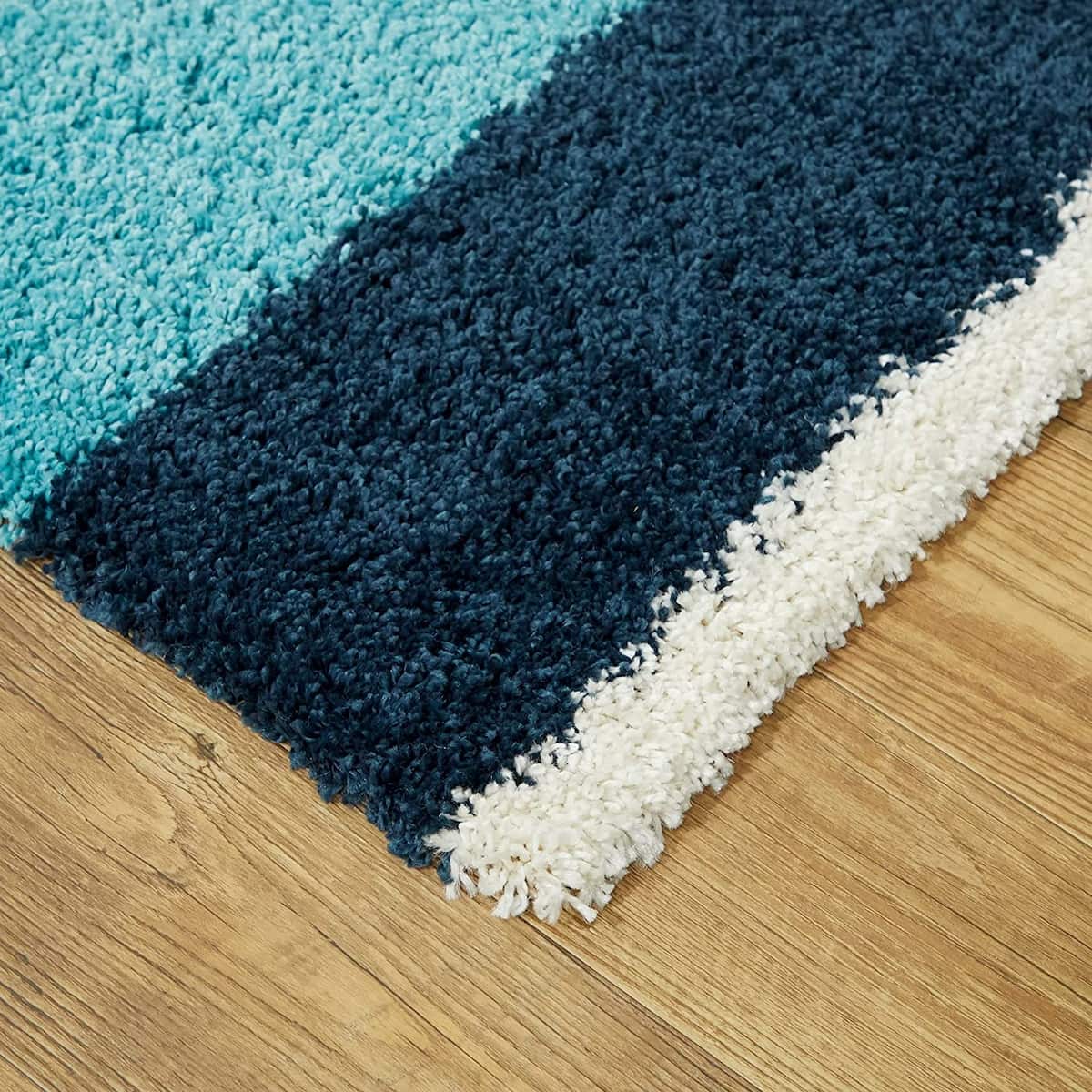A close-up of a plush shag rug with diagonal stripes in turquoise, dark navy, and off-white, shown on a light brown wood floor.
