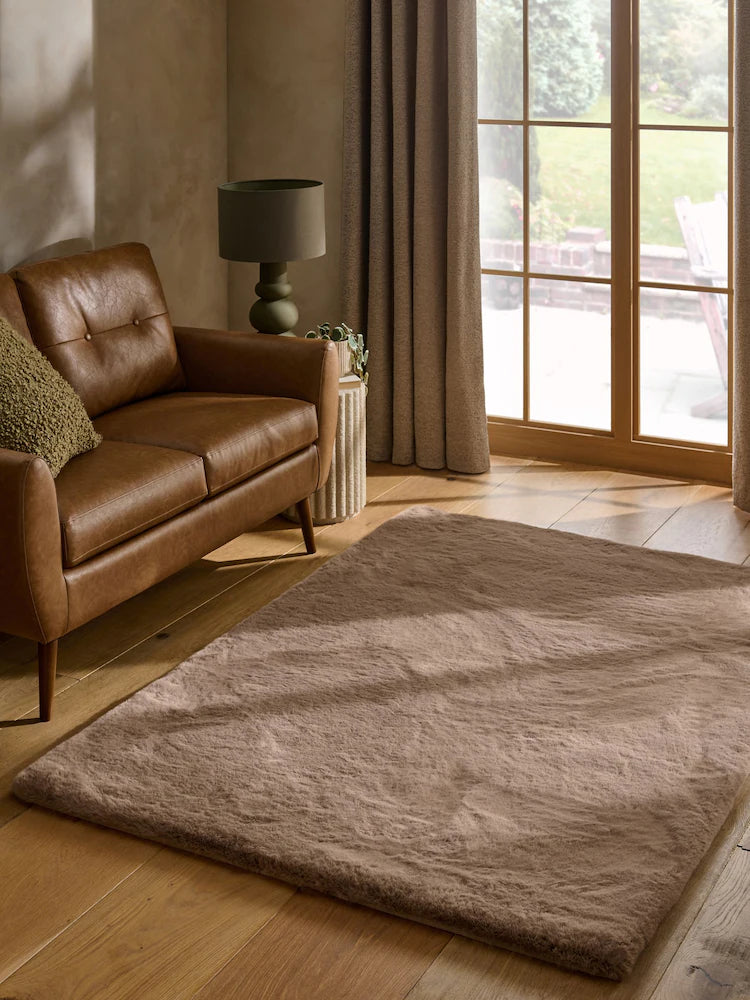 A rectangular shag area rug in a warm taupe beige color, styled in a modern living room next to a brown leather sofa and a large window.