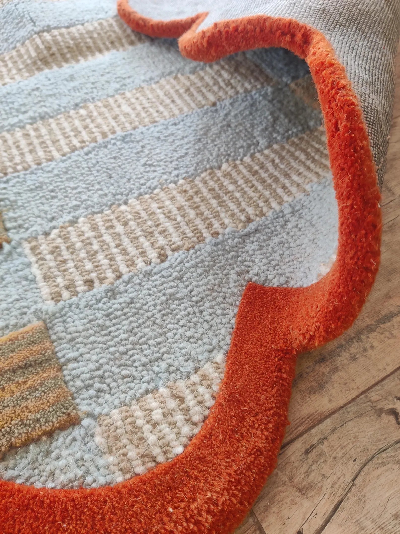 A close-up of a textured area rug with horizontal stripes in light blue, beige, and cream, featuring a thick, plush burnt orange border, laid on a rustic wooden floor.