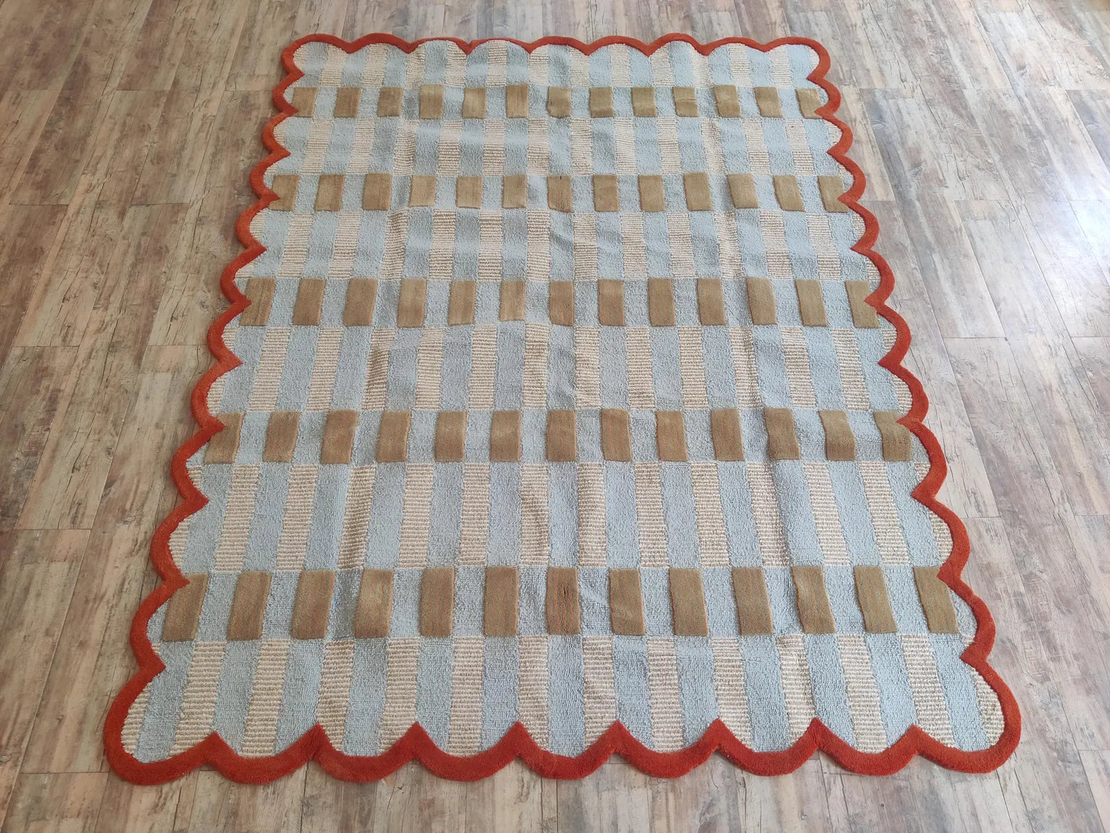 A rectangular area rug with a scalloped border, featuring a woven pattern of beige and light blue vertical stripes with horizontal bands of brown, laid out on a wood-look floor.