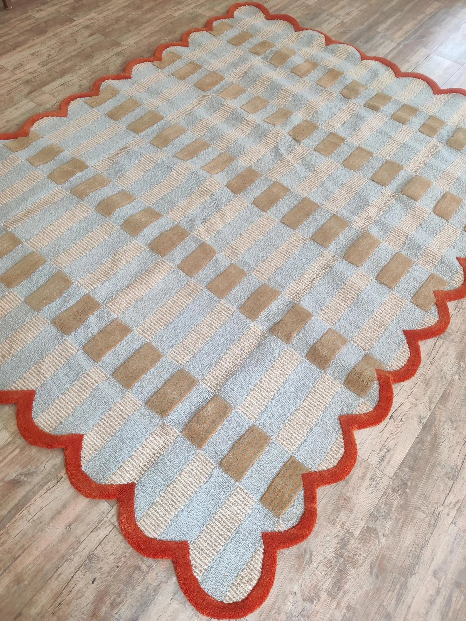 A rectangular area rug with a scalloped edge, featuring a woven basketweave pattern in light blue, beige, and brown, finished with a rust-red border, shown on a wooden floor.