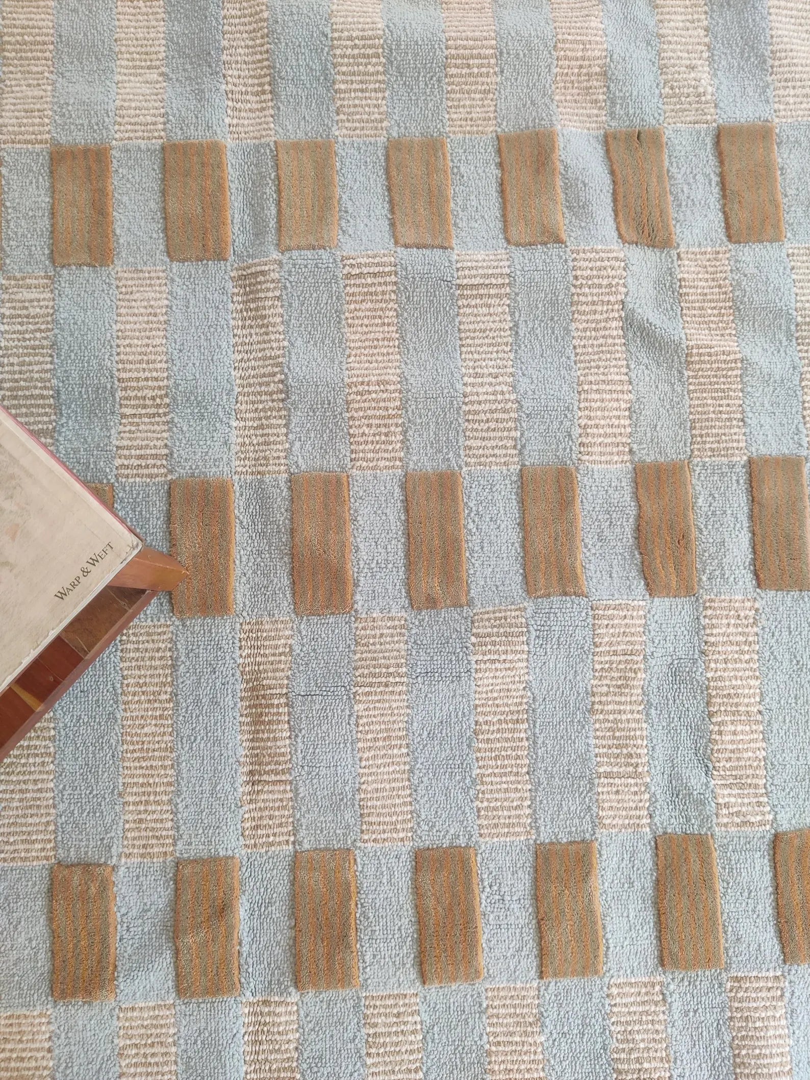 A close-up of a woven textile with a checkered pattern in light blue, beige, and tan, with a small portion of a book visible in the lower-left corner.