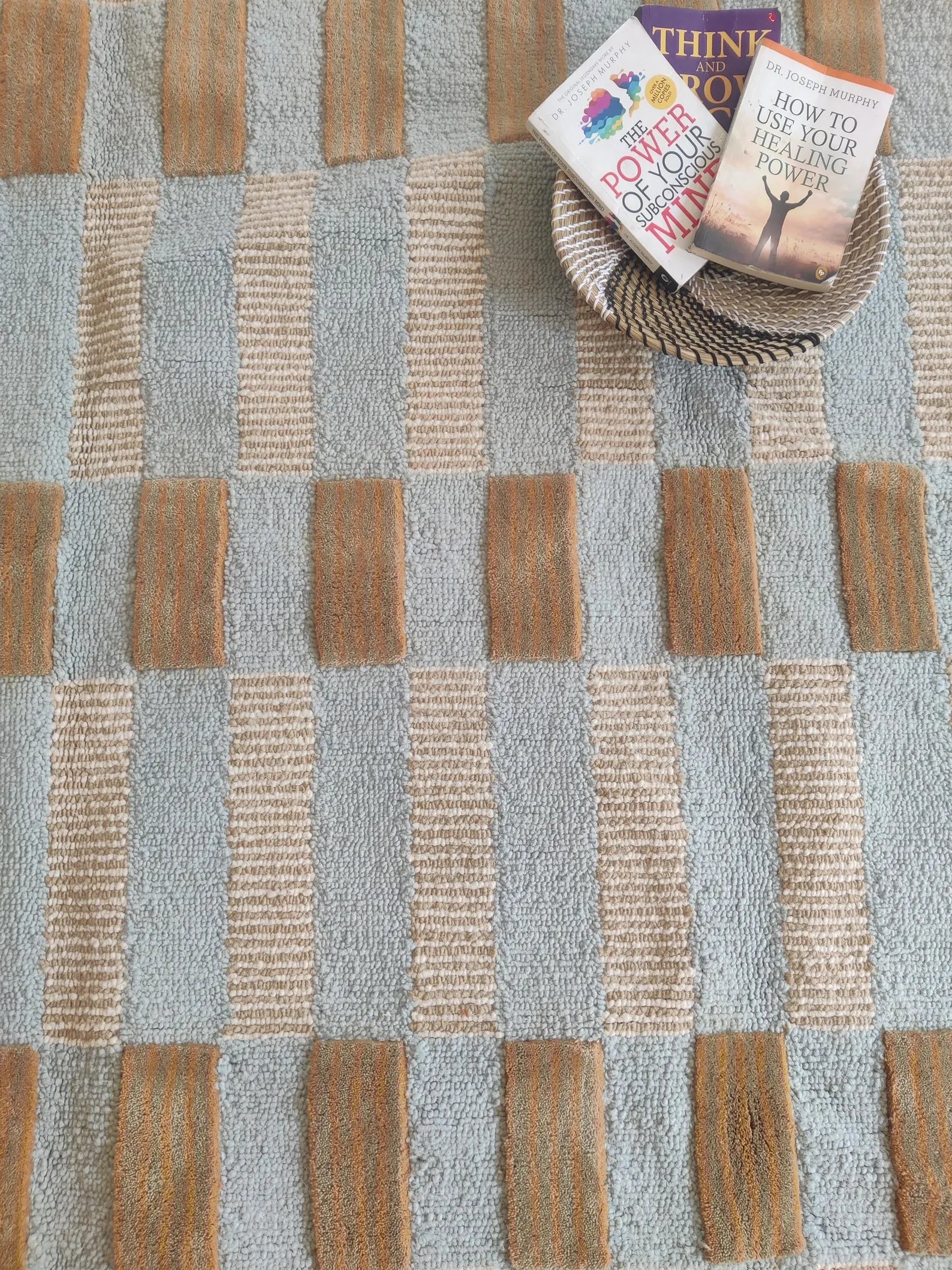 A close-up of a woven area rug with a geometric pattern of light blue-gray and natural tan stripes, styled with two books on a small woven tray in the upper right corner.
