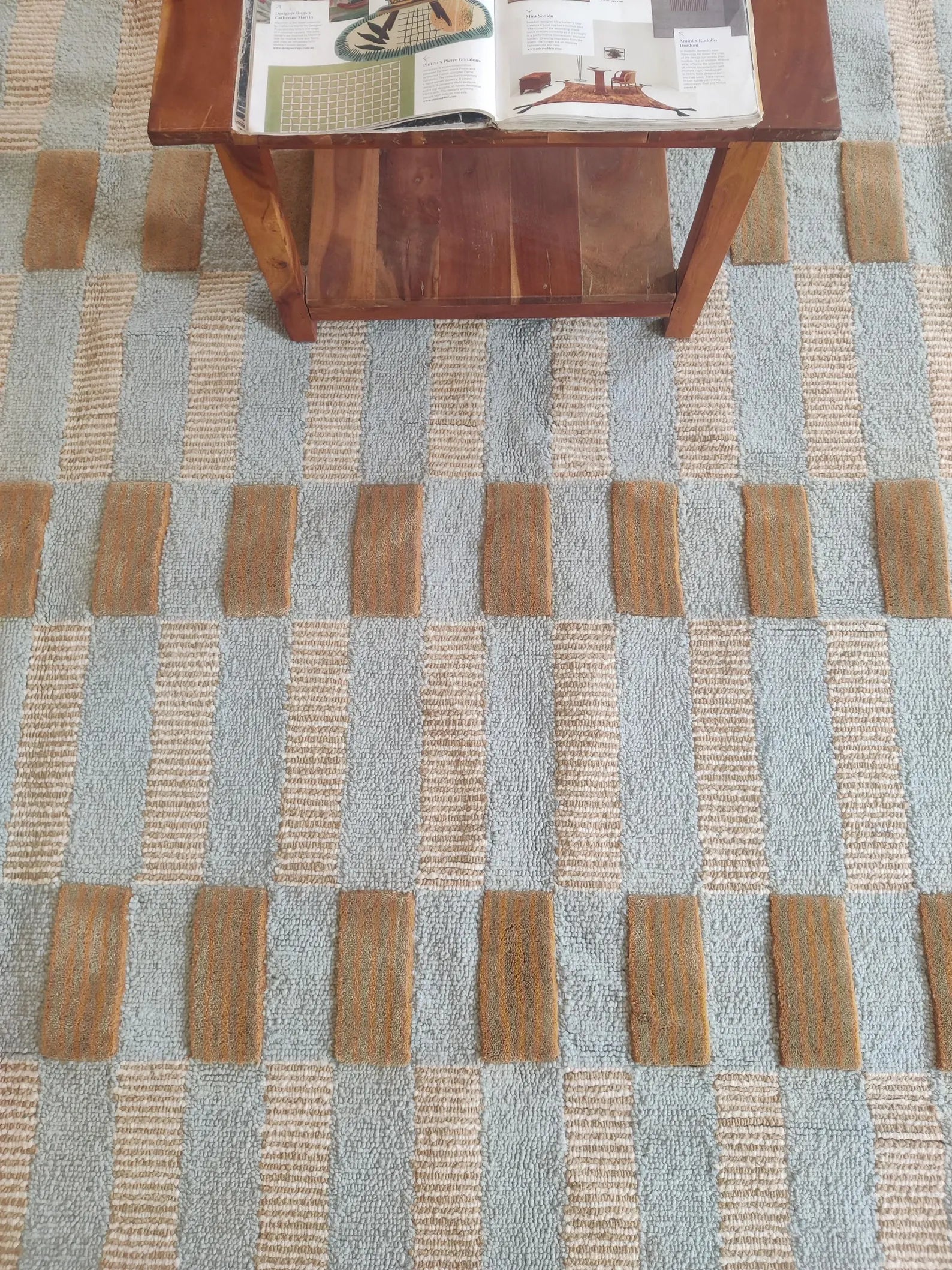 A close-up of a woven area rug with a geometric checkered pattern in light blue-gray and natural beige, with a wooden table partially visible at the top.