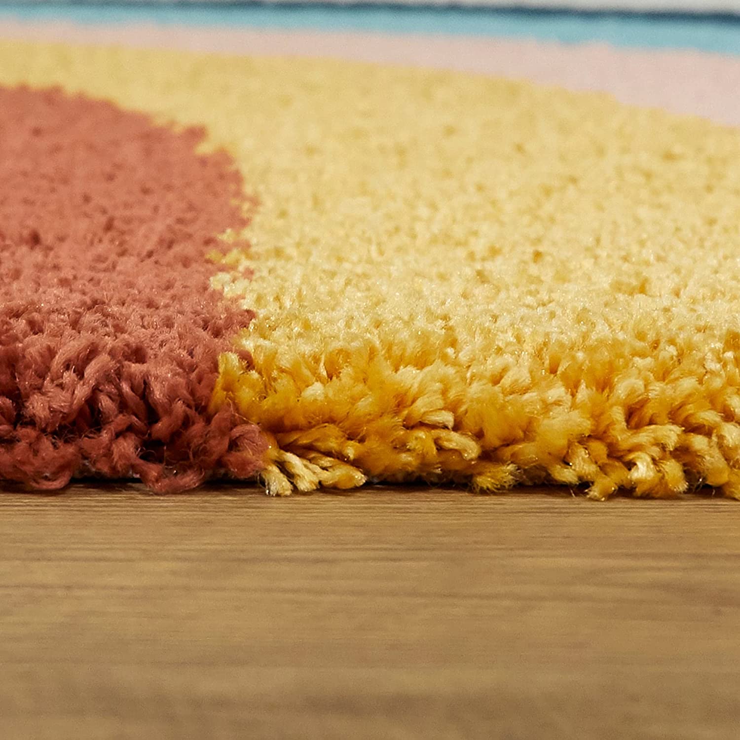 Close-up of a plush shag rug with a wavy color-blocked pattern, showing a dusty rose section on the left, a large pale yellow section in the center, and a hint of a light blue section at the top, resting on a wooden floor.