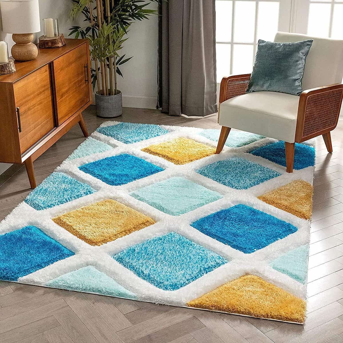 A RugNoor's colorful geometric patterned microfiber and silk rug in blue, white, and yellow, placed on a wooden floor under a chair and a potted plant.