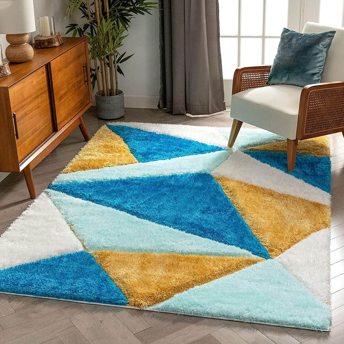A RugNoor's geometric patterned rug with blue, white, and yellow colors, placed in a room with wooden furniture and a gray curtain.