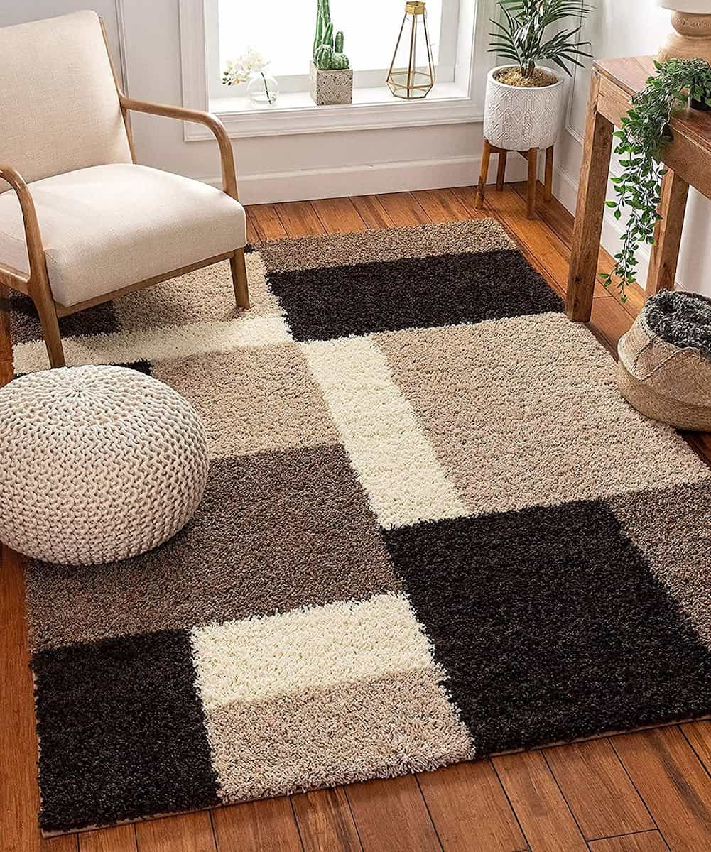 RugNoor's shaggy rug with a geometric pattern in black, beige, and brown colors, placed on a wooden floor under a white chair and a potted plant.