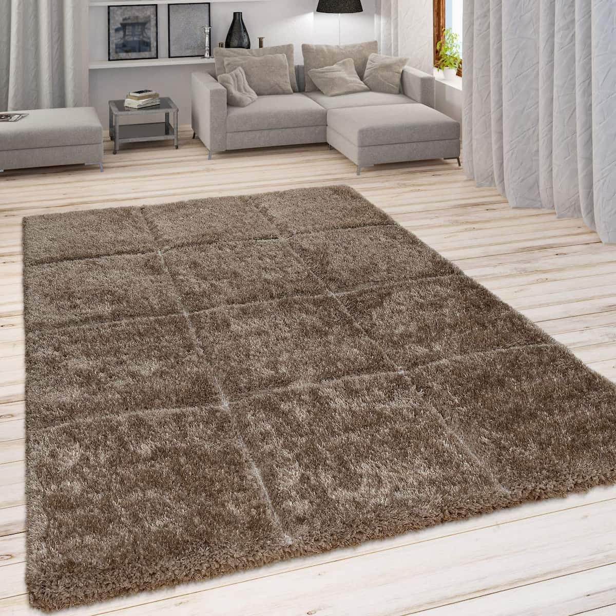 A RugNoor's brown fluffy area rug placed on a wooden floor in a living room setting with a couch and armchairs in the background.