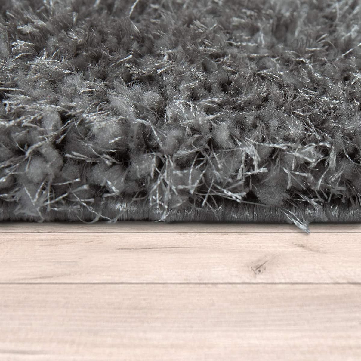 RugNoor's Close-up of a dark gray shaggy rug with a thick pile, shown from below at the edge of a light wood floor.