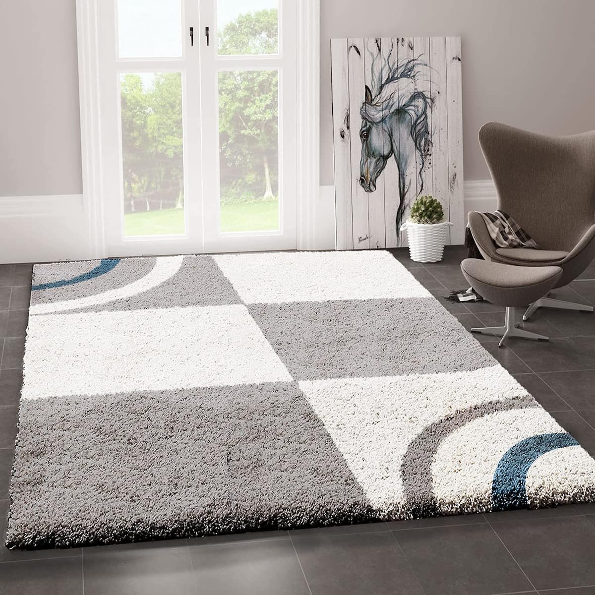 RugNoor's modern shaggy rug with a geometric pattern in gray, white, and blue colors, placed on a tiled floor in a room with a chair and a framed picture.