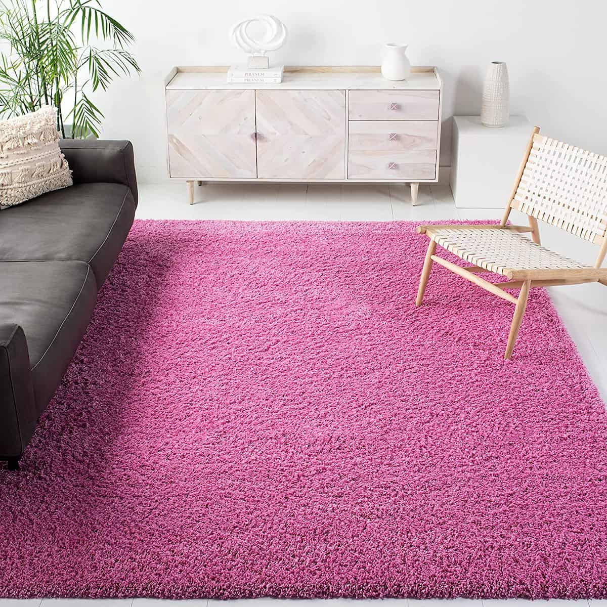 A pink shaggy rug laid out on the floor beside a black sofa and a wooden chair in a room with a white coffee table and plant decor - RugNoor