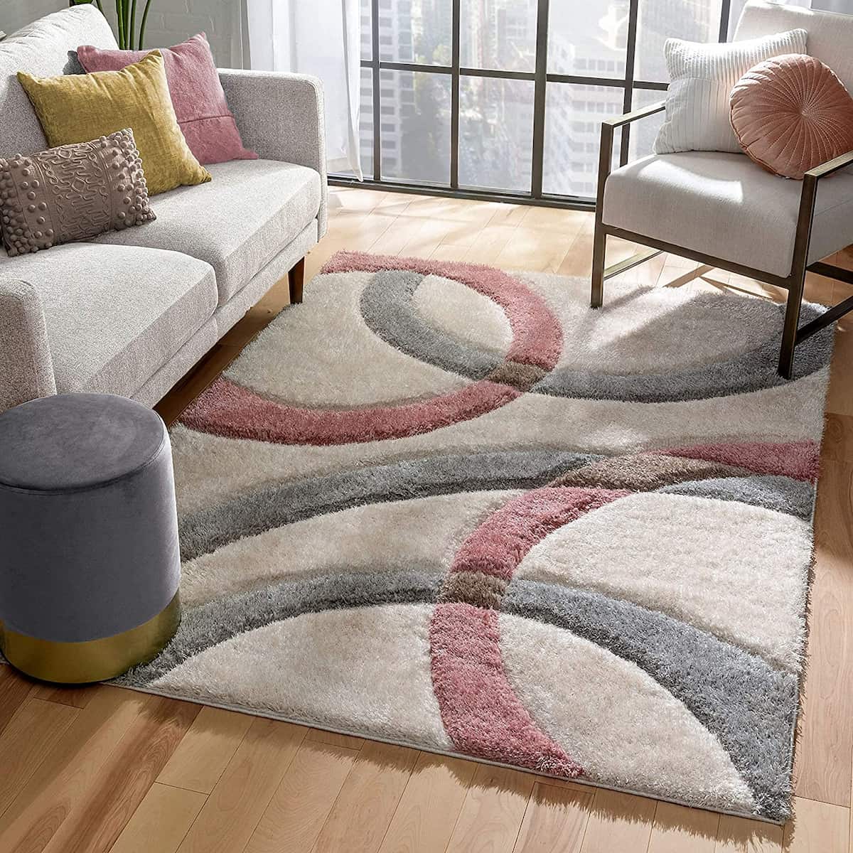 A RugNoor's modern living room setting with a pink, gray, and white fluffy rug on wooden flooring. The rug has a geometric pattern, and it is accompanied by a white sofa, a armchair with pink cushion, and a gray ottoman.