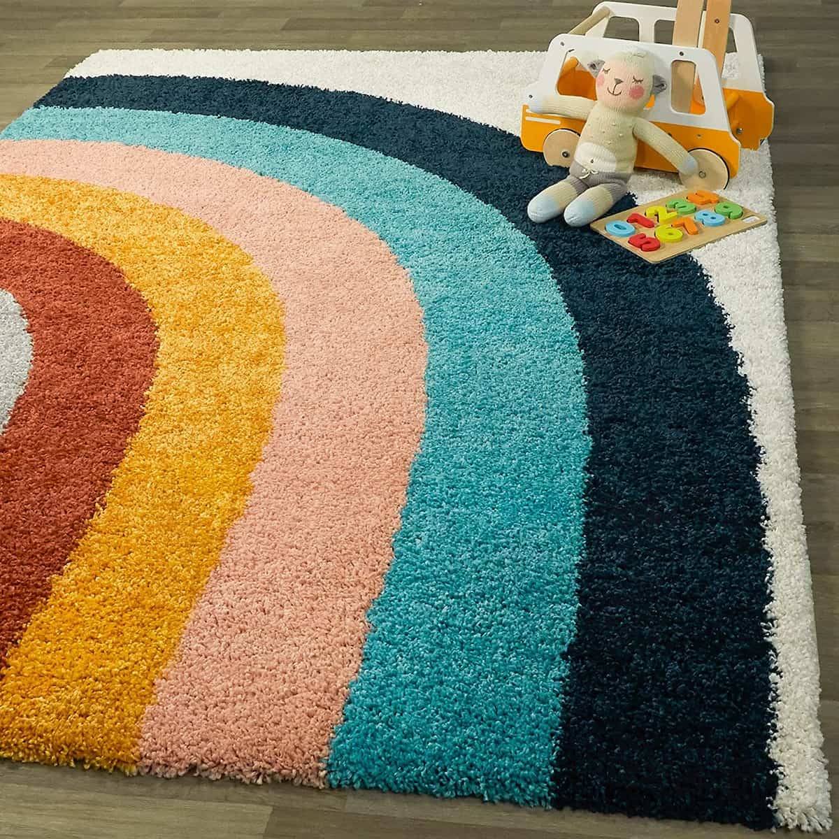 RugNoor's multicolored microfiber shaggy rug with a semi-circular pattern in shades of white, blue, navy, pink, gold, red, and grey, placed on a wooden floor with a toy car and a doll on top of it.