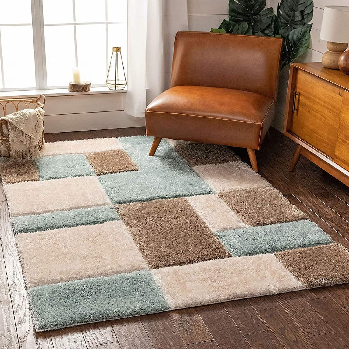 A RugNoor shaggy rug with a block-shaped design in blue, cream, and brown colors, placed on a wooden floor under a leather armchair and a wooden side table with a plant.